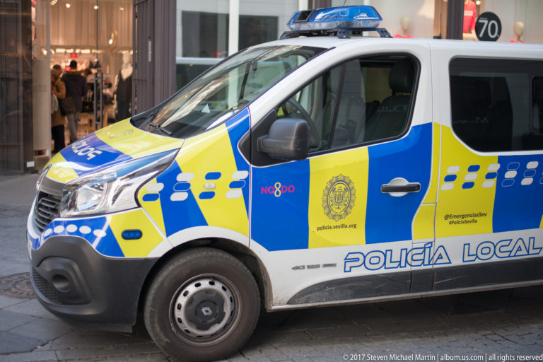 White Seville Policía Local van with blue-and-yellow markings and roof light bar parked along a city street.