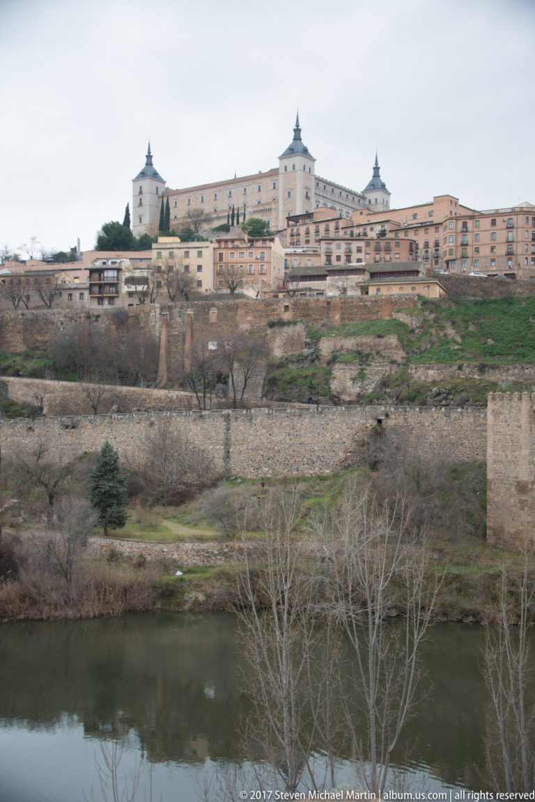 Alcázar of Toledo on a high ridge above the Tagus River, with stone walls and sloping ground in the foreground.
