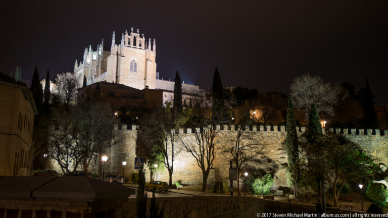 Illuminated historic complex above stone walls and trees on a hillside in Toledo at night.