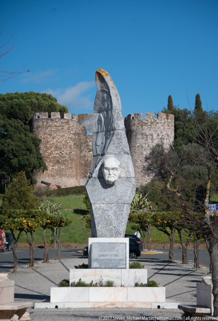 Angular gray stone memorial with carved figure and projecting emblem, backed by crenellated castle walls and small citrus trees.