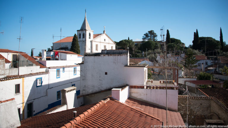 White church and narrow bell tower rising above red-tiled roofs, garden walls and cypress trees in Vila Viçosa.