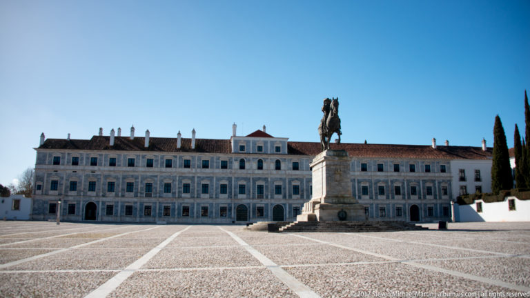 Long marble façade of the Ducal Palace across a paved square, with a large equestrian statue on a stone pedestal in front.