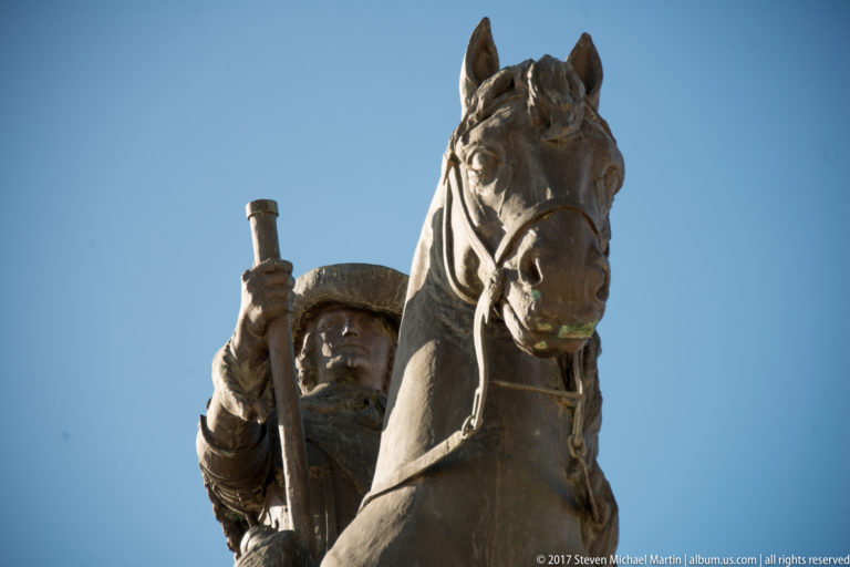 Close upward view of a bronze mounted ruler holding a baton, with the horse’s head prominent against blue sky.