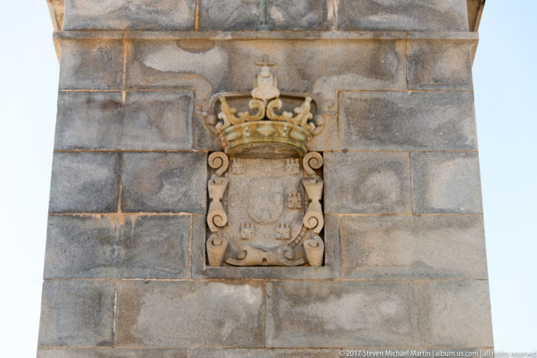 Carved Portuguese shield beneath a large royal crown and scrollwork on a pale stone monument pedestal.