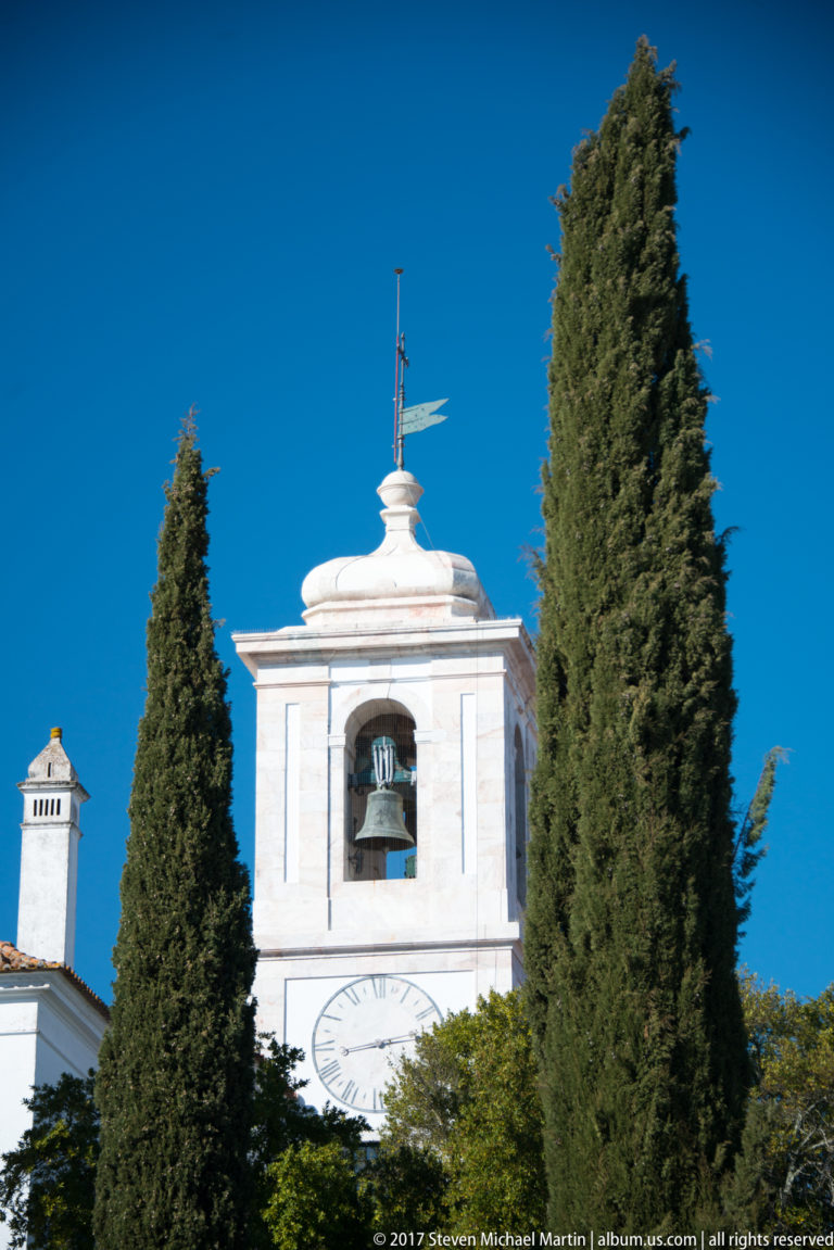 White square bell tower with one visible bell and a clock face, framed by two tall cypress trees.