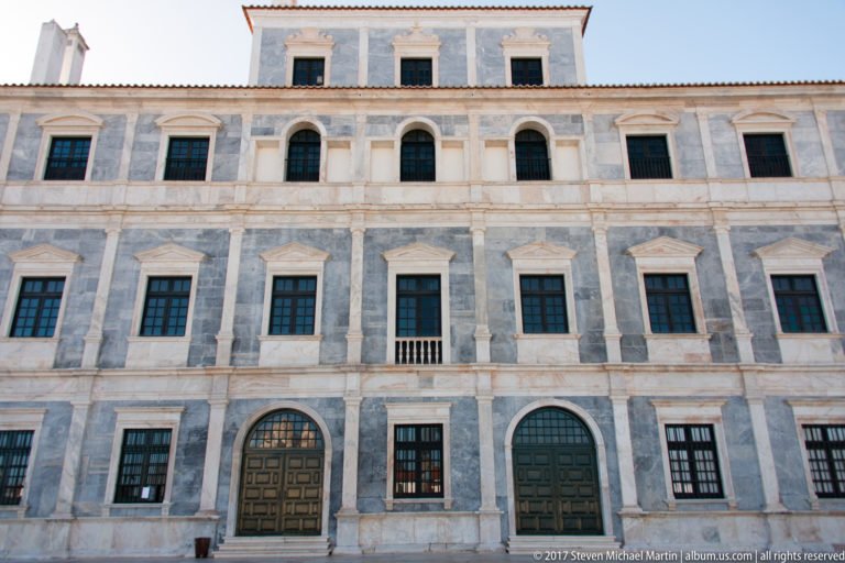 Close upward view of the Ducal Palace’s marble façade with rows of framed windows and two large dark wooden doorways.