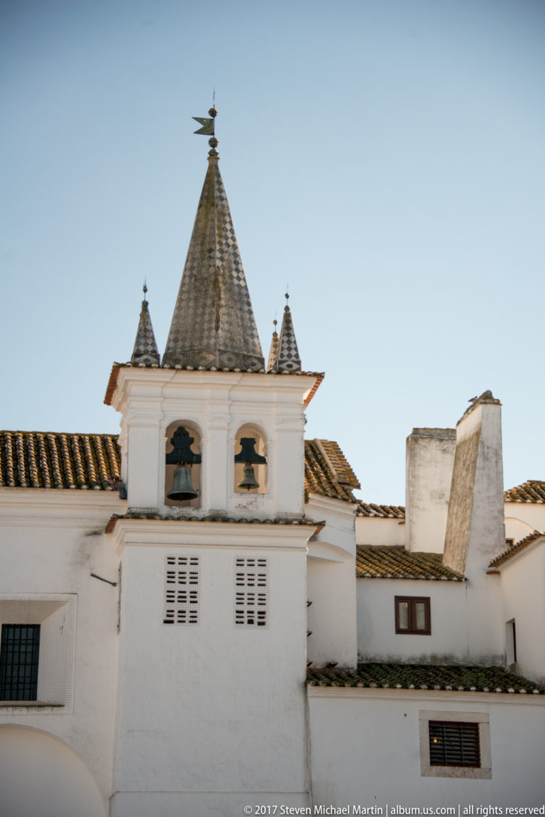 White rooftop turret with two bells, a steep pyramidal roof, corner pinnacles and a weather vane.