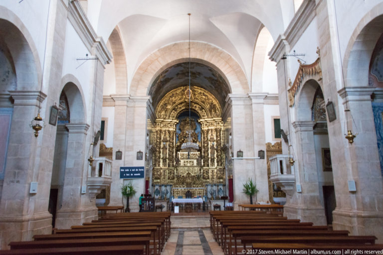 Church nave with wooden pews and pale stone arches leading to a richly gilded high altar.