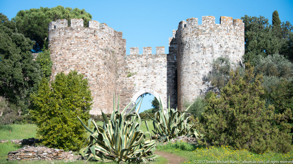 Arched castle entrance between two round crenellated stone towers, with agaves and shrubs in the foreground.