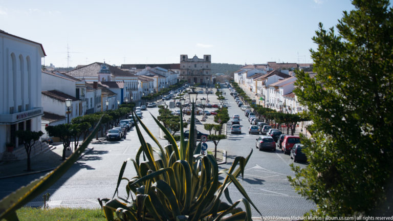 Long tree-lined avenue through whitewashed Vila Viçosa, viewed from above with cars and a distant landmark centered on the horizon.