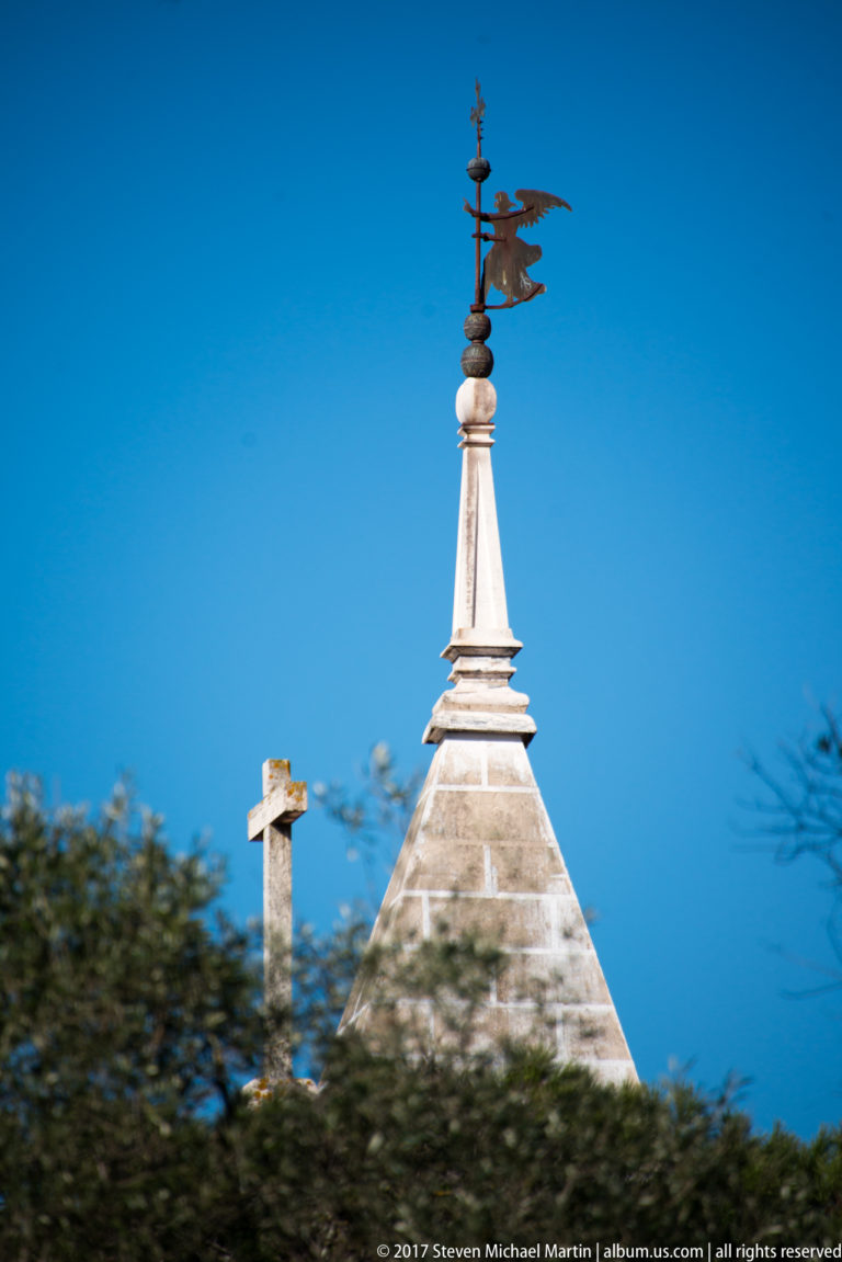 Metal weather vane on a layered stone finial above a pyramidal roof, with a stone cross nearby against blue sky.