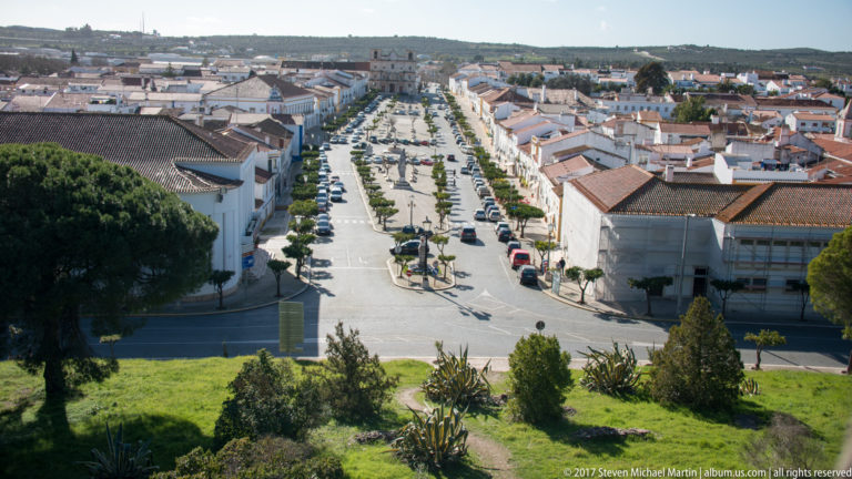 Elevated panorama of Vila Viçosa with a long landscaped avenue, white buildings, red roofs and low hills beyond.