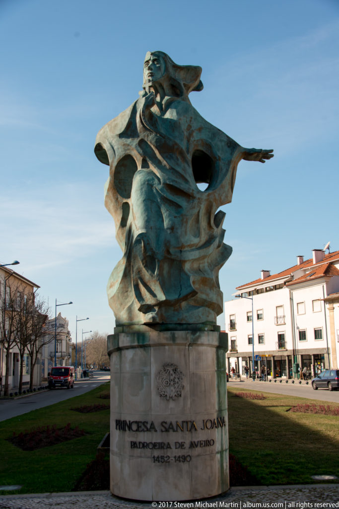 Bronze monument to Princess Saint Joana on a stone pedestal in an Aveiro square under a clear blue sky.