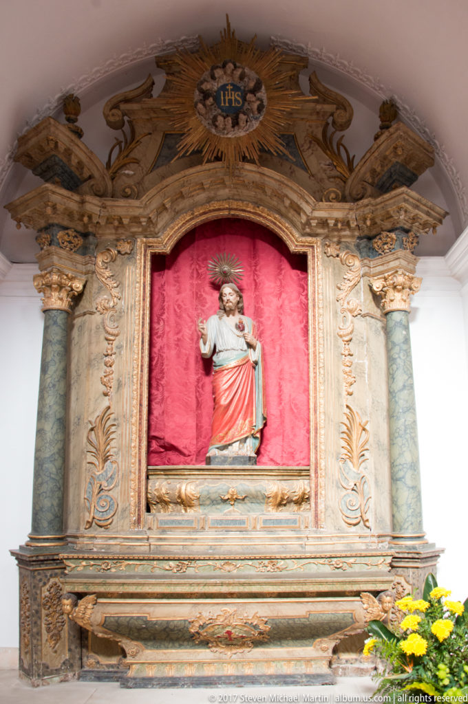 Standing Christ figure in a small ornate retable with red background, blue-green columns, gilded carving and an IHS emblem above.