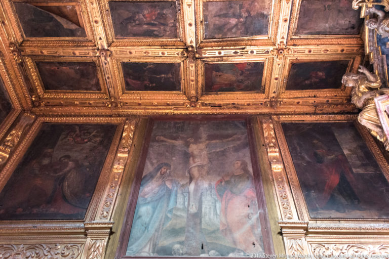Upward view of a painted coffered ceiling meeting large framed religious paintings divided by gilded carved moldings.