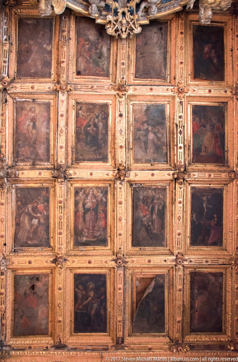 Upward view of a ceiling arranged as a grid of dark narrative paintings within carved and gilded rectangular coffers.