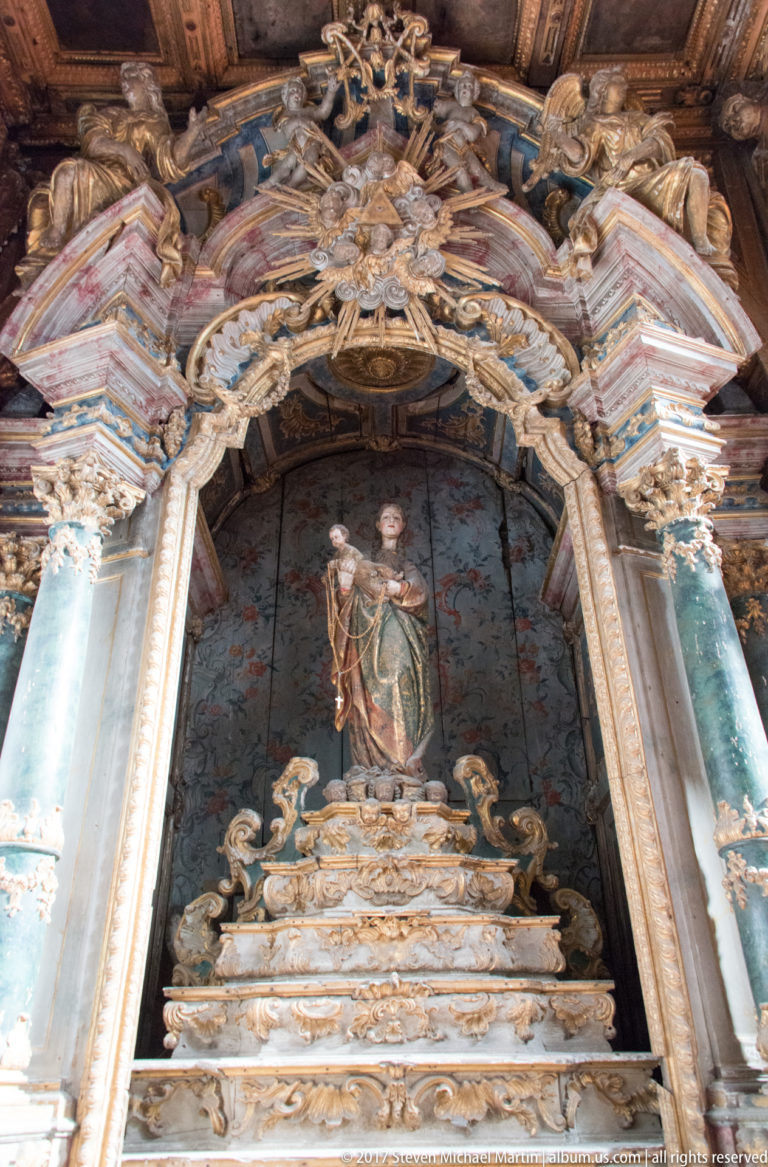 Standing female saint in an ornate niche framed by blue-green columns, gilded scrollwork and a deeply carved canopy.