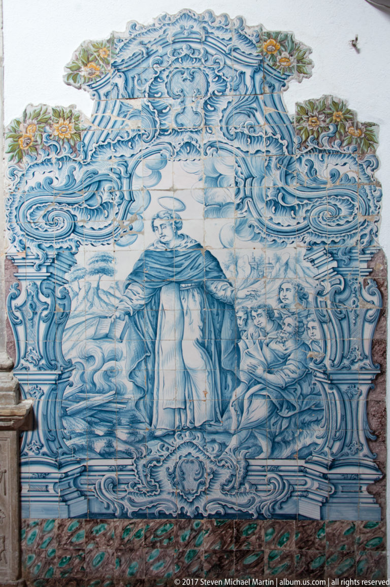 Blue-and-white tiled mural with a robed central figure, surrounding people and an elaborate painted ornamental border.