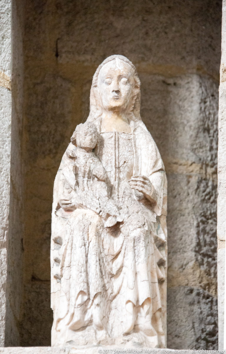 Weathered pale stone sculpture of a frontal female religious figure with deeply carved drapery, displayed against a dark wall.