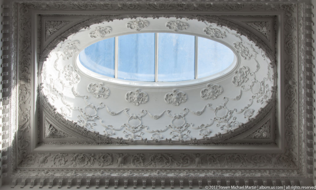 Oval skylight in a white ornamental ceiling framed by plaster garlands, floral rosettes and layered molded borders.