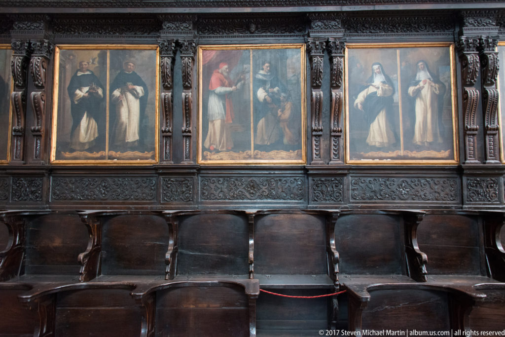 Dark wooden choir stalls beneath three framed religious paintings, with carved pilasters and decorative panels surrounding the ensemble.