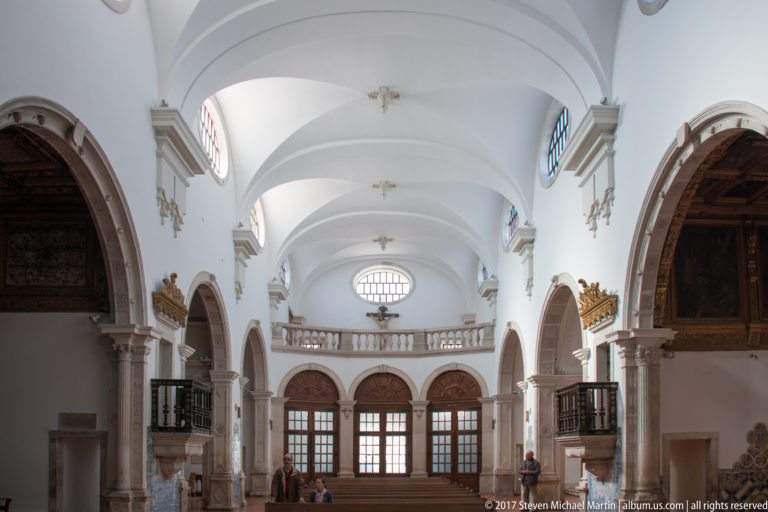 Symmetrical white-vaulted interior with repeated side arches, dark wooden furnishings, high windows and a balustraded gallery at the far end.