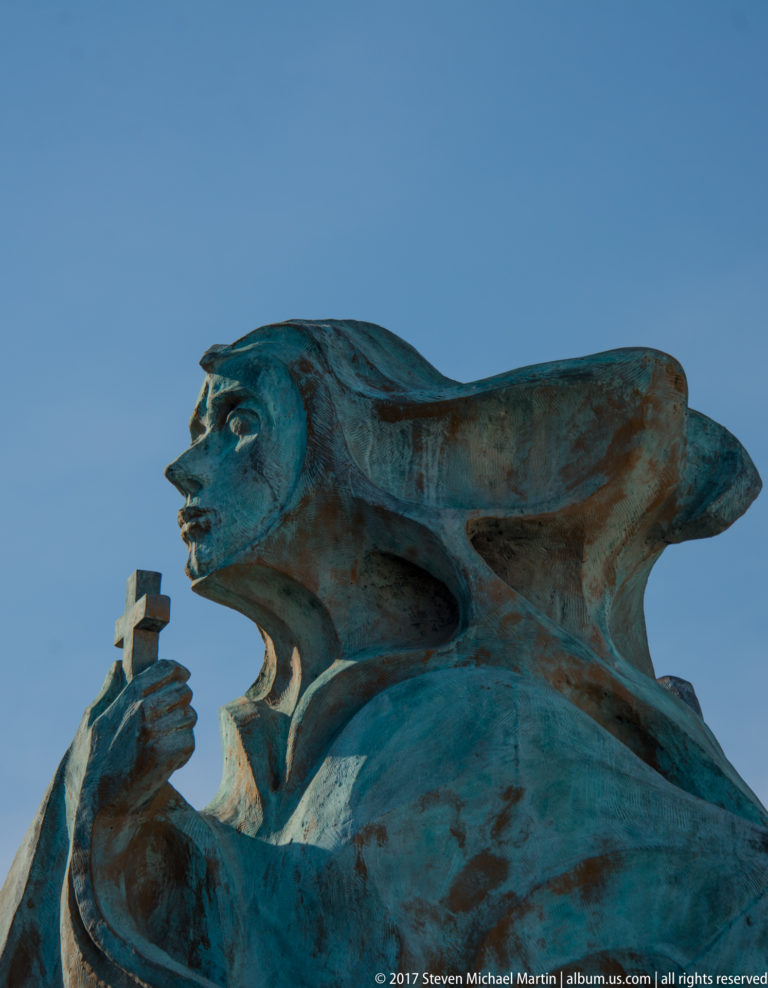 Low-angle close view of a patinated bronze female figure holding a small cross, with wind-swept drapery against blue sky.