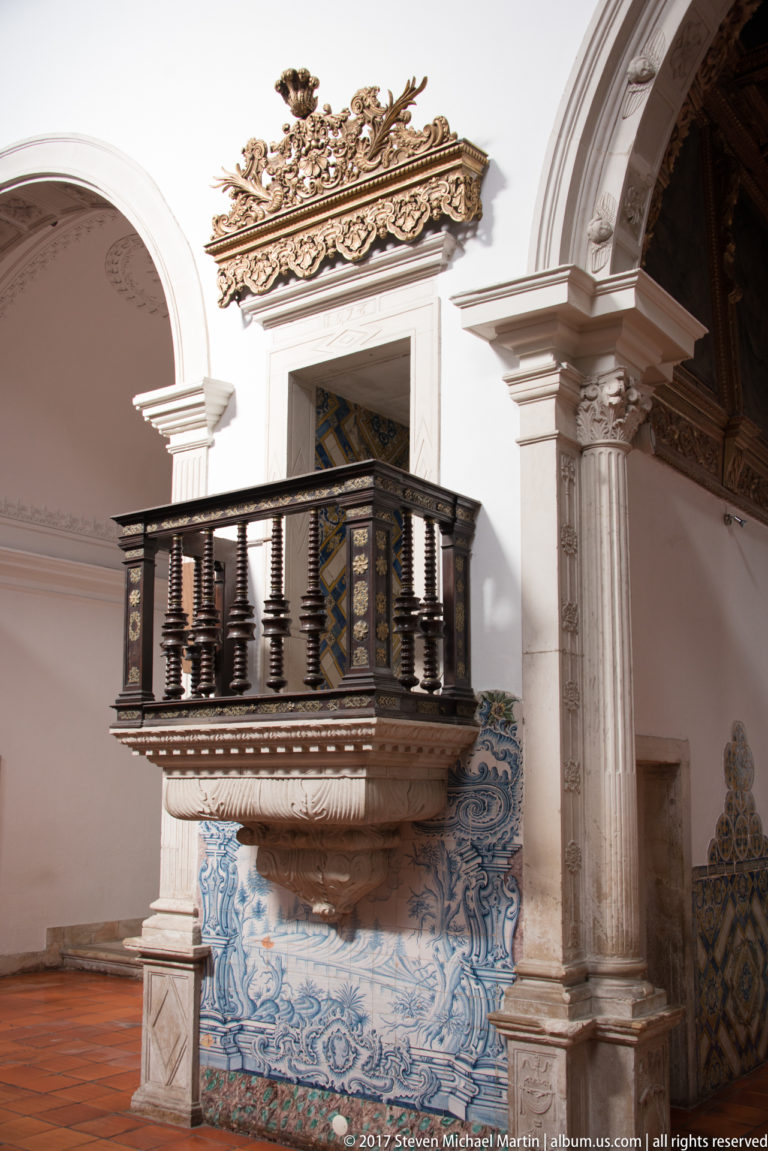 Dark wooden balcony with turned balusters above blue-and-white tiles, beside a large pale stone arch and column.