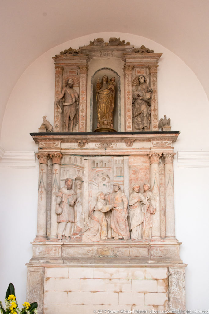 Pale carved stone monument with a crowded narrative relief below and three sculptural figures arranged across the upper register.