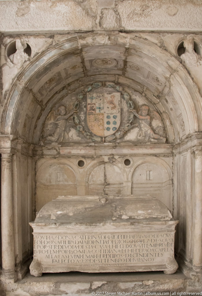 Inscribed rectangular stone tomb set beneath a shallow carved arch, with a heraldic shield centered above the recess.