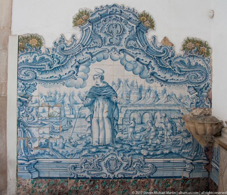 Blue-and-white tiled mural with a robed figure before sailing ships and waterfront activity, bordered by elaborate scrollwork.