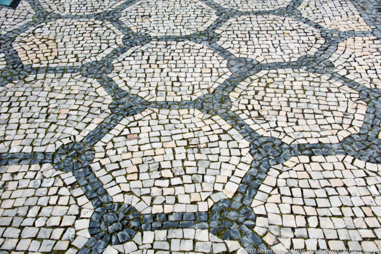 Close downward view of Portuguese black-and-white cobblestone pavement arranged in repeating circles and interlocking dark bands.