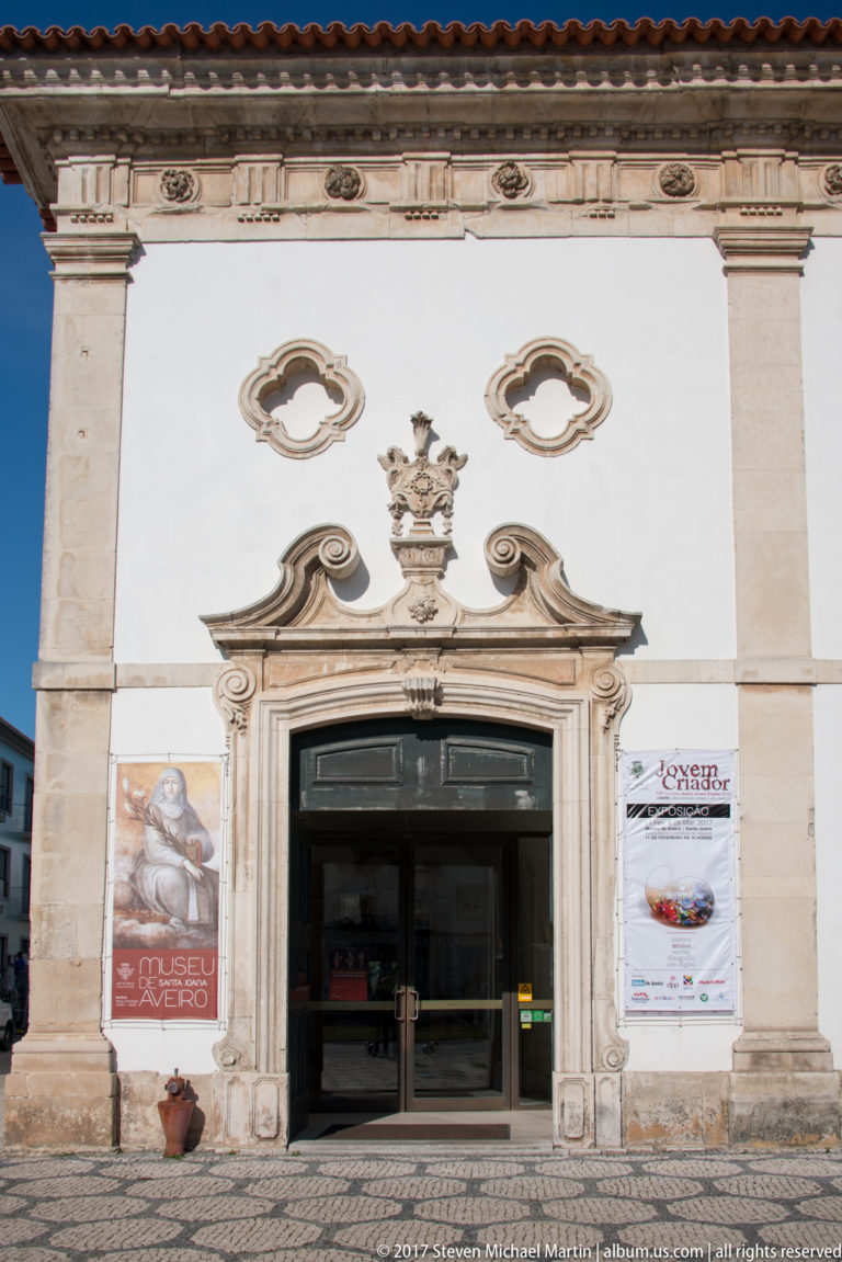 White historic façade with ornate carved stone doorway, two oval windows and exhibition posters on either side of the entrance.