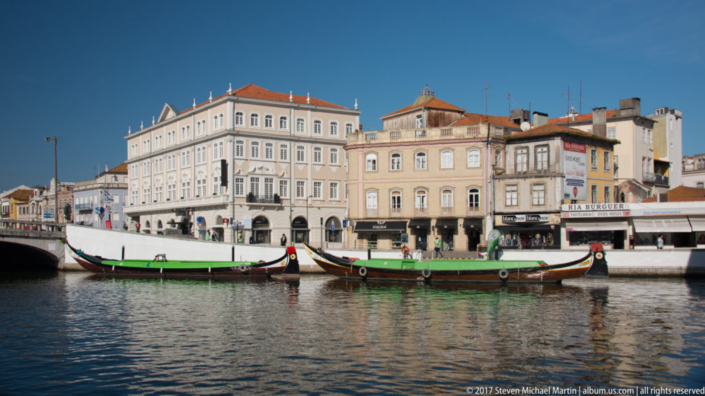 Several long painted moliceiro boats moored on an Aveiro canal in front of multi-story historic buildings under blue sky.