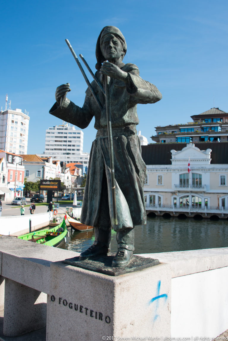 Bronze statue labeled O Fogueteiro, showing a traditionally dressed figure carrying long implements beside an Aveiro canal.