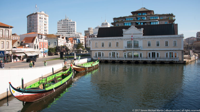 Painted moliceiro boats moored along an Aveiro canal, facing a white waterfront building with city towers rising behind it.