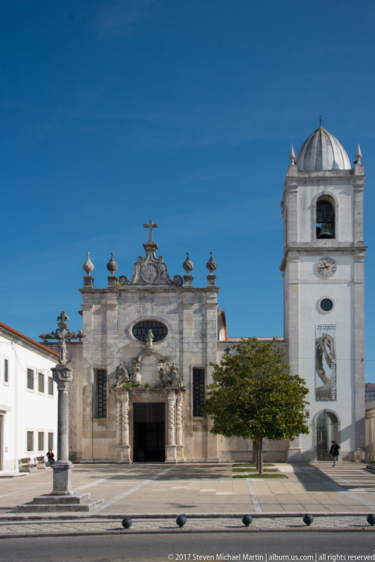 Frontal view of an ornate stone church façade beside a white clock and bell tower in a paved square.