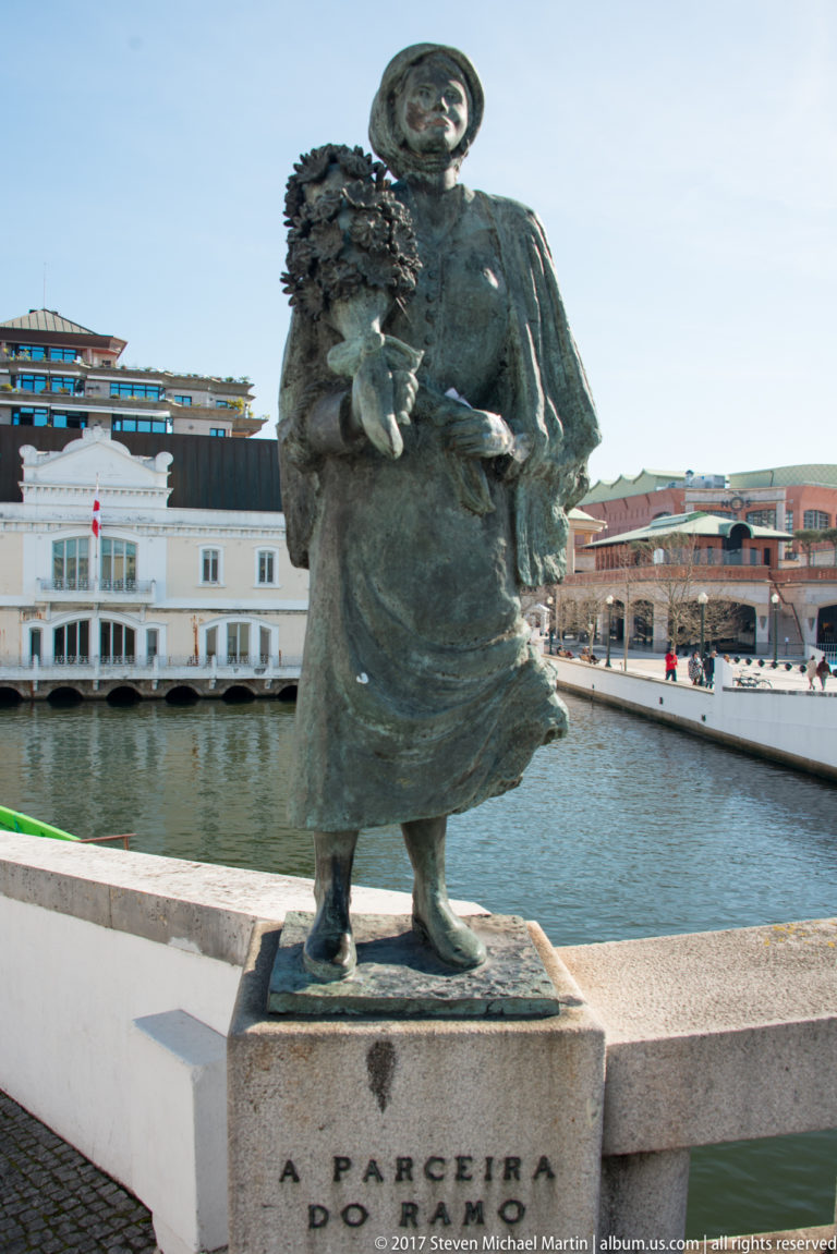 Bronze statue labeled A Parceira do Ramo, depicting a traditionally dressed woman carrying a leafy bundle beside the canal.