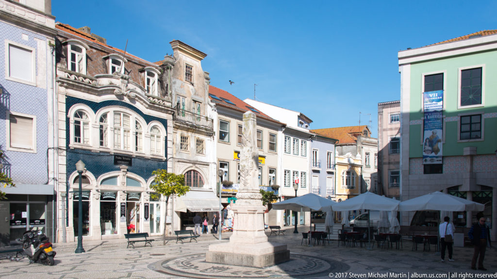 Ornate multi-story façade beside a stone monument, café umbrellas, pedestrians, and patterned pavement in an Aveiro square.