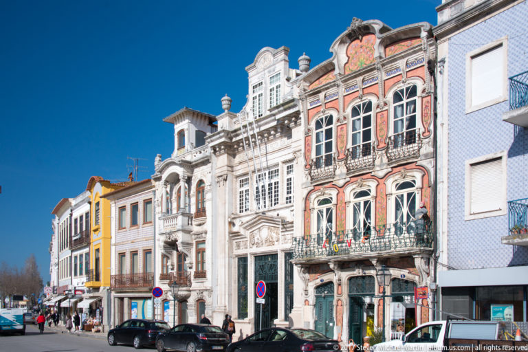 Row of ornate white and pink townhouses with arched windows, wrought-iron balconies, cars, and pedestrians under a blue sky.