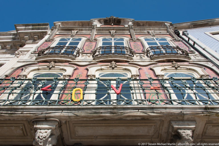 Low-angle view of a pink ornate façade with arched windows, carved details, and stacked wrought-iron balconies.