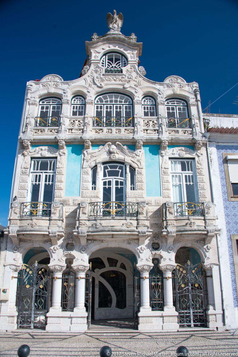 Symmetrical Art Nouveau façade with blue-green panels, pale carved stone, arched openings, decorative metalwork, and a sculpted roofline.