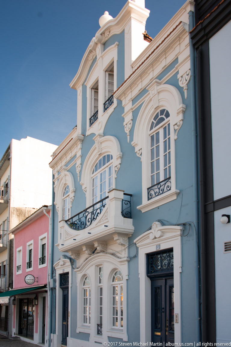Pale blue townhouse with white curved ornament, tall windows, small iron balconies, and a shaped decorative roofline.