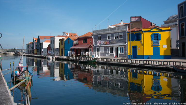 Colorful waterfront buildings, including a yellow-and-blue façade, reflected in a calm Aveiro canal beside a narrow quay.