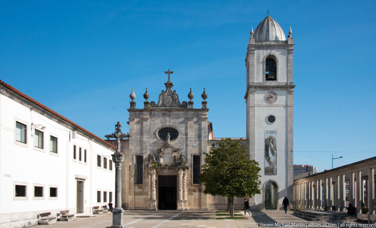 Oblique view across a paved forecourt toward an ornate stone church façade and adjacent white bell tower.