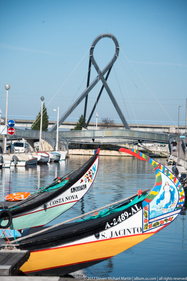 Colorfully painted moliceiro prows in the foreground with a modern looping pedestrian bridge spanning the canal behind them.