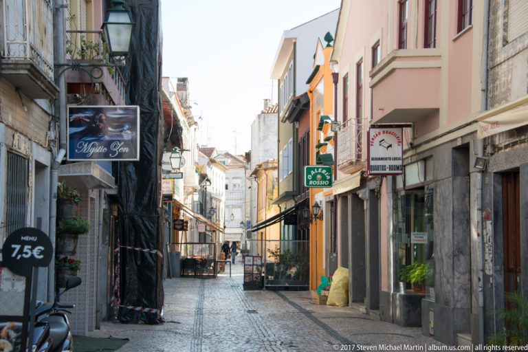 Narrow paved street lined with small shops, restaurant signs, awnings, café furniture, and closely spaced upper-story façades.