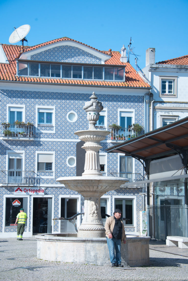 Carved stone fountain in front of a blue-and-white tiled building with rows of windows and ground-floor shops.
