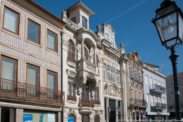 Oblique street view of ornate tiled and carved façades with balconies, arched windows, colorful surfaces, and a black streetlamp.