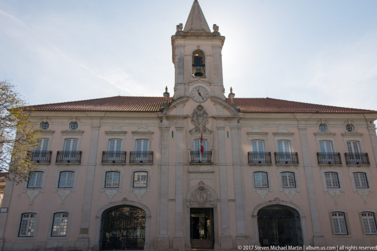 Frontal view of Aveiro City Hall, a pale-pink civic building with a central clock tower, iron balconies, and arched entrances.